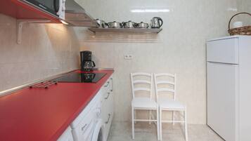 Een kleine keuken met een rood aanrecht, een koffiezetapparaat, een magnetron en een witte koelkast. Er staan twee witte stoelen en boven het aanrecht bevindt zich een plank met keukengerei.