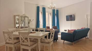 A dining area with a white table and chairs, a blue sofa, a TV mounted on the wall, and a chandelier.