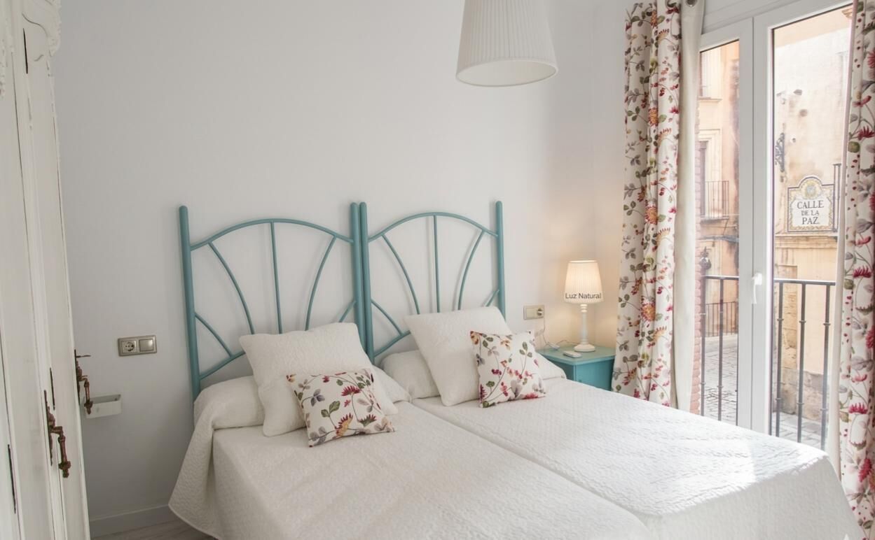 A bed with a white bedspread and floral pillows, a teal headboard, a nightstand with a lamp, and a view of a street through a window with floral curtains.