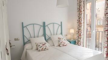 A bed with a white bedspread and floral pillows, a teal headboard, a nightstand with a lamp, and a view of a street through a window with floral curtains.