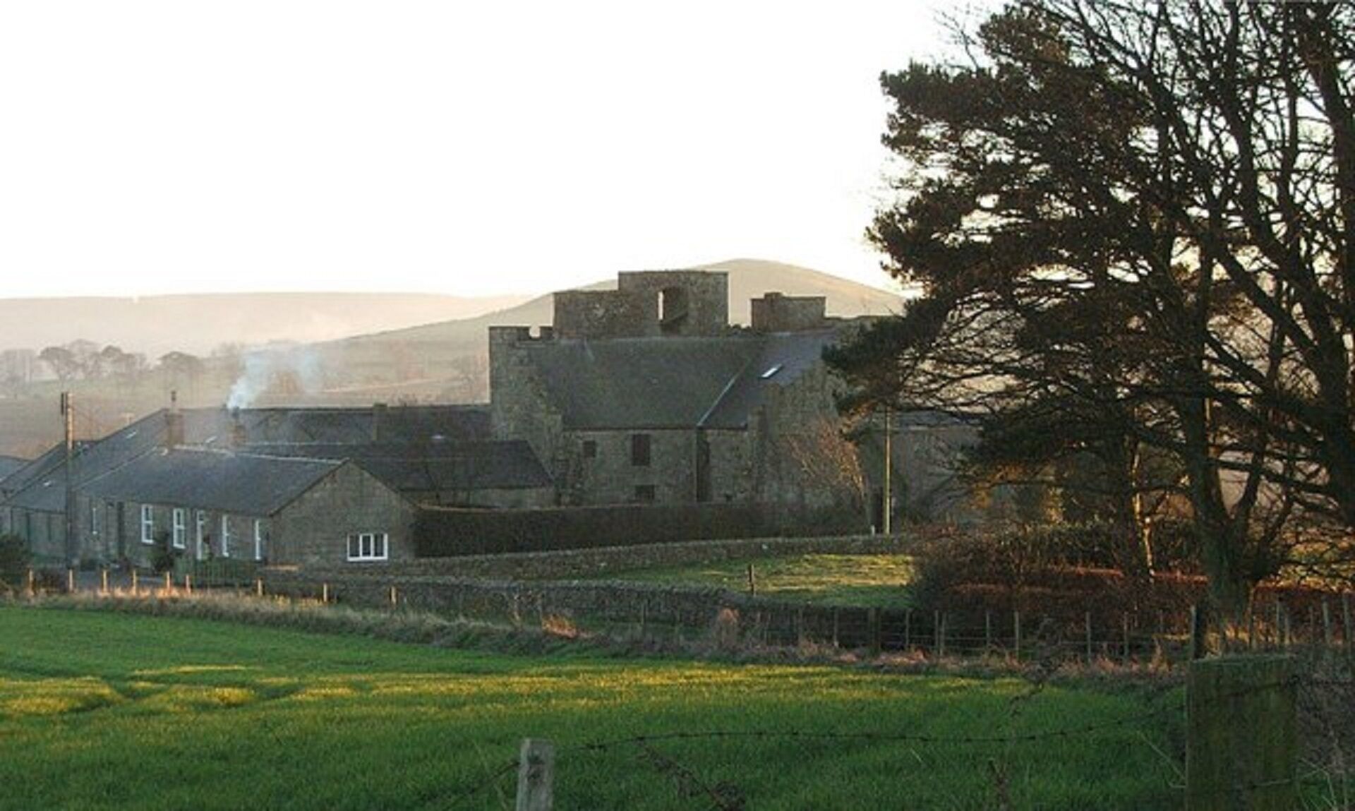 Ein Steinhaus mit einem markanten Turm, umgeben von ländlicher Landschaft mit Feldern und einem Baum.