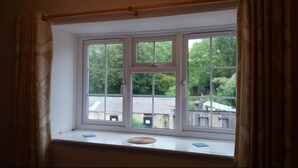 A window with a view of a residential area, curtains, and a small shelf with items. - 'The Stables'. A lovely stone built cottage in the small village of Powburn. (Powburn)