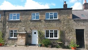 A stone building with a white door, multiple windows, and a small garden area with potted plants and outdoor furniture. - 'The Stables'. A lovely stone built cottage in the small village of Powburn. (Powburn)