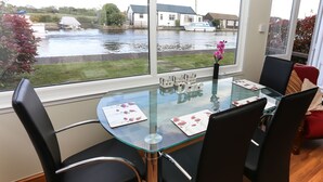 Uma área de refeições com mesa de vidro, cadeiras pretas e vista para um canal com barcos.