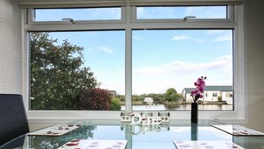 Um quarto com uma mesa de vidro e duas cadeiras, uma janela com vista para um canal e edifícios, e um vaso com flores sobre a mesa.
