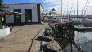 Una plaza frente al mar con un edificio identificado como "Plaza de la Marina" y una barrera de seguridad cerca del agua.