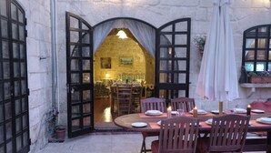 A dining area with a stone wall, a wooden table, and chairs. There is a doorway leading to a kitchen with a hanging light. - Id-Dwejra  House  of Character with Privite Pool,  swim Jet and Jaccuzzi (Zejtun)