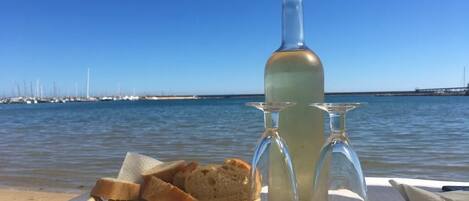 A table with a bottle of wine, two wine glasses, and a basket of bread on a beach with a view of the sea and wind turbines in the background.