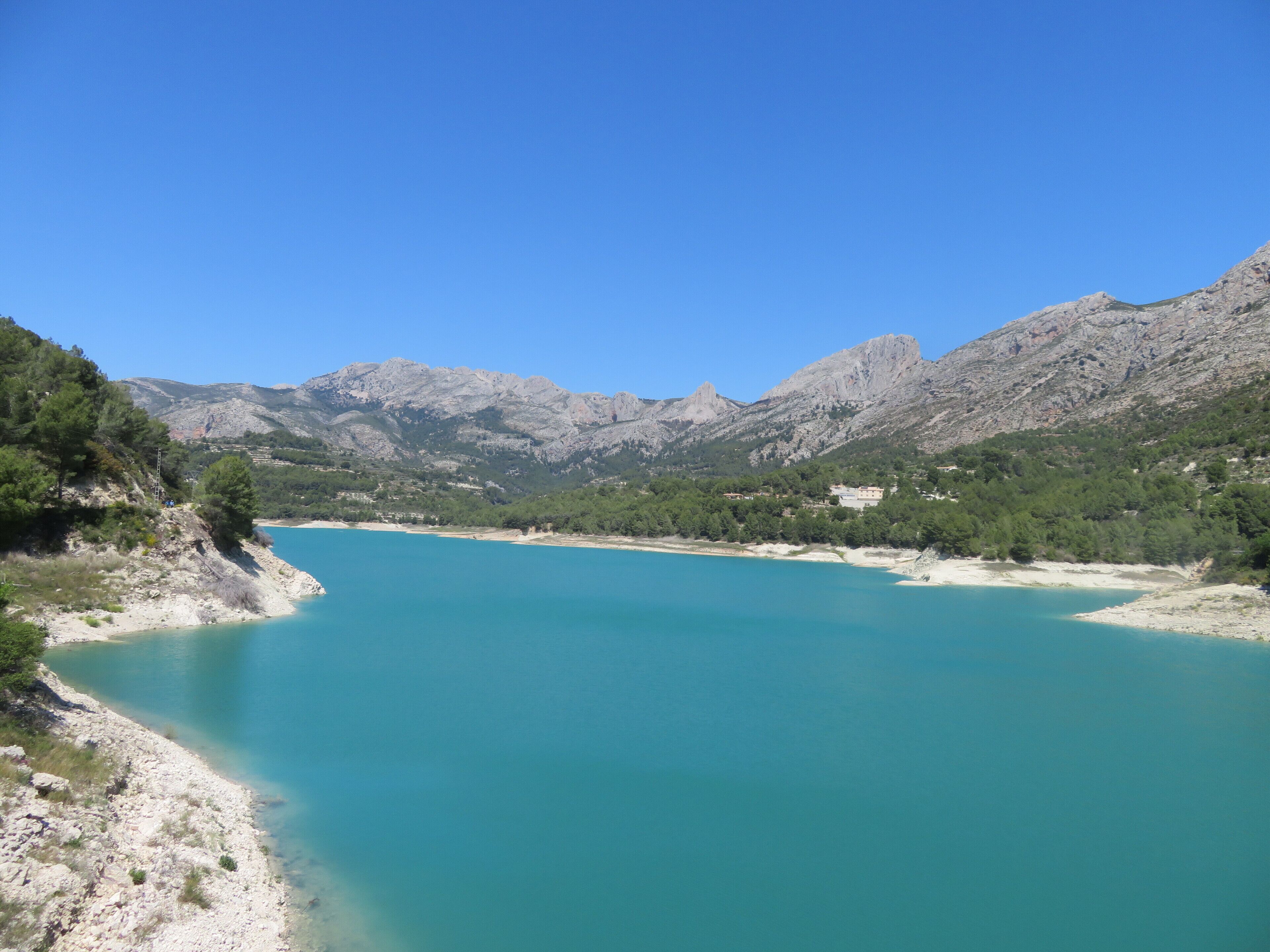 A coastal landscape with a turquoise lake, rocky mountains, and sparse vegetation.