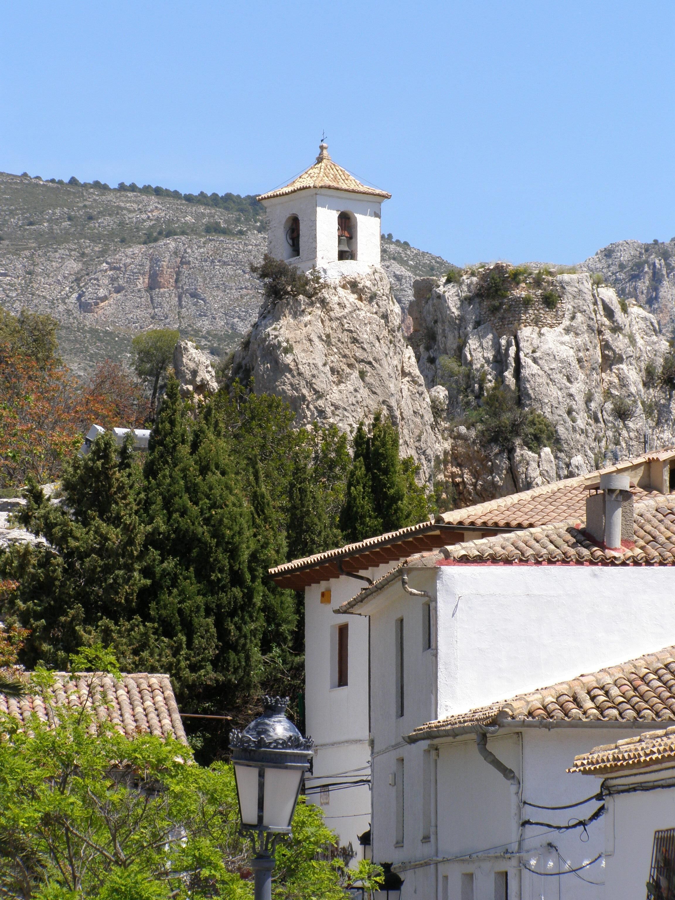 Un bâtiment doté d’un clocher, situé sur une colline rocheuse et entouré d’arbres et d’autres constructions.