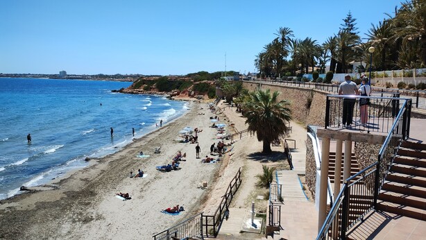 Una playa con gente tomando el sol y nadando, una escalera que baja a la playa, y una vista despejada del océano.