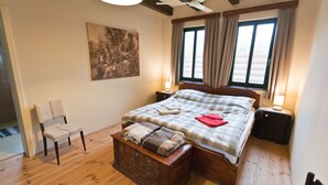 Ein Schlafzimmer mit Holzboden, einem Bett mit kariertem Überwurf, einem Sessel, einem Nachttisch, einer Lampe und einem Fenster mit Vorhängen.