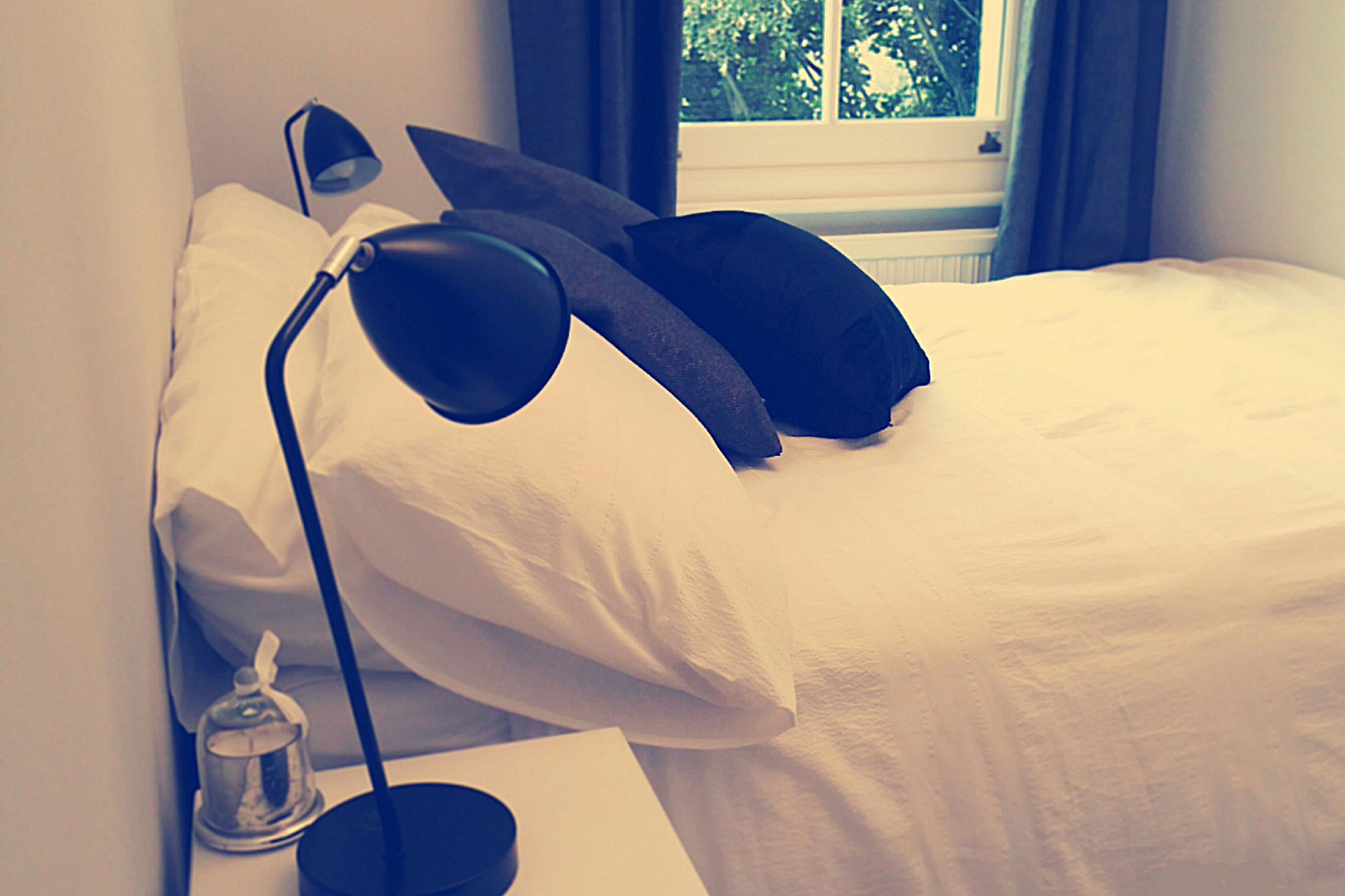 En pænt redt seng med hvide sengetøj, en sort lampe og et vindue med gardiner.