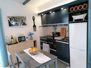 A modern kitchen with a microwave, toaster, and coffee maker on the counter. There is a bowl of fruit on the table and a bottle of wine. The kitchen has dark cabinets and a white refrigerator.
