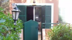 Degraus externos levam a uma porta com persianas verdes, em meio à vegetação e a um poste de luz.