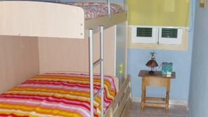 Uma cama beliche com colcha listrada e colorida, um pequeno criado-mudo de madeira com uma luminária e uma janela com cortinas amarelas.