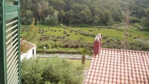 Vista de um telhado de telhas vermelhas, um edifício branco e uma paisagem verde com árvores e uma colina ao fundo.