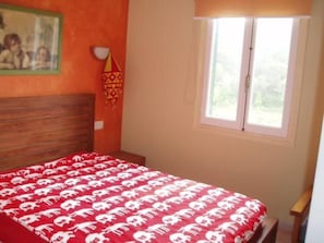 A bedroom with a wooden headboard, a window with white frames, and a red bedspread with white patterns.