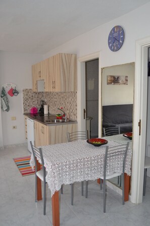 A compact kitchen with a dining area, including a table with a lace tablecloth, chairs, and a clock on the wall. - Sun & Sol apartment in Caleta de Fuste (Castillo Caleta de Fuste)
