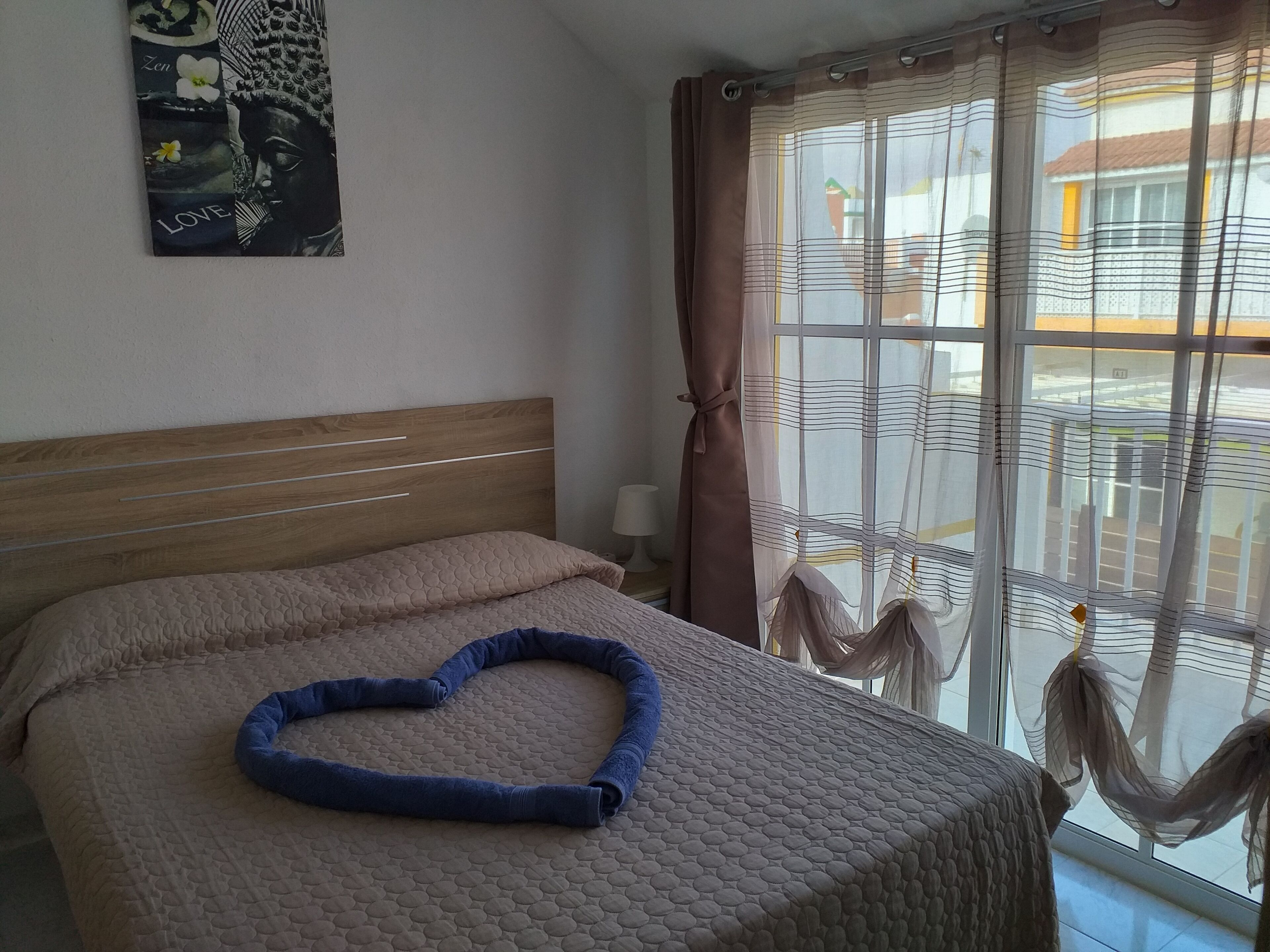 Ein Schlafzimmer mit einem Bett, einem herzförmigen Kissen, einem Fenster mit Vorhängen und einem Wandbild.