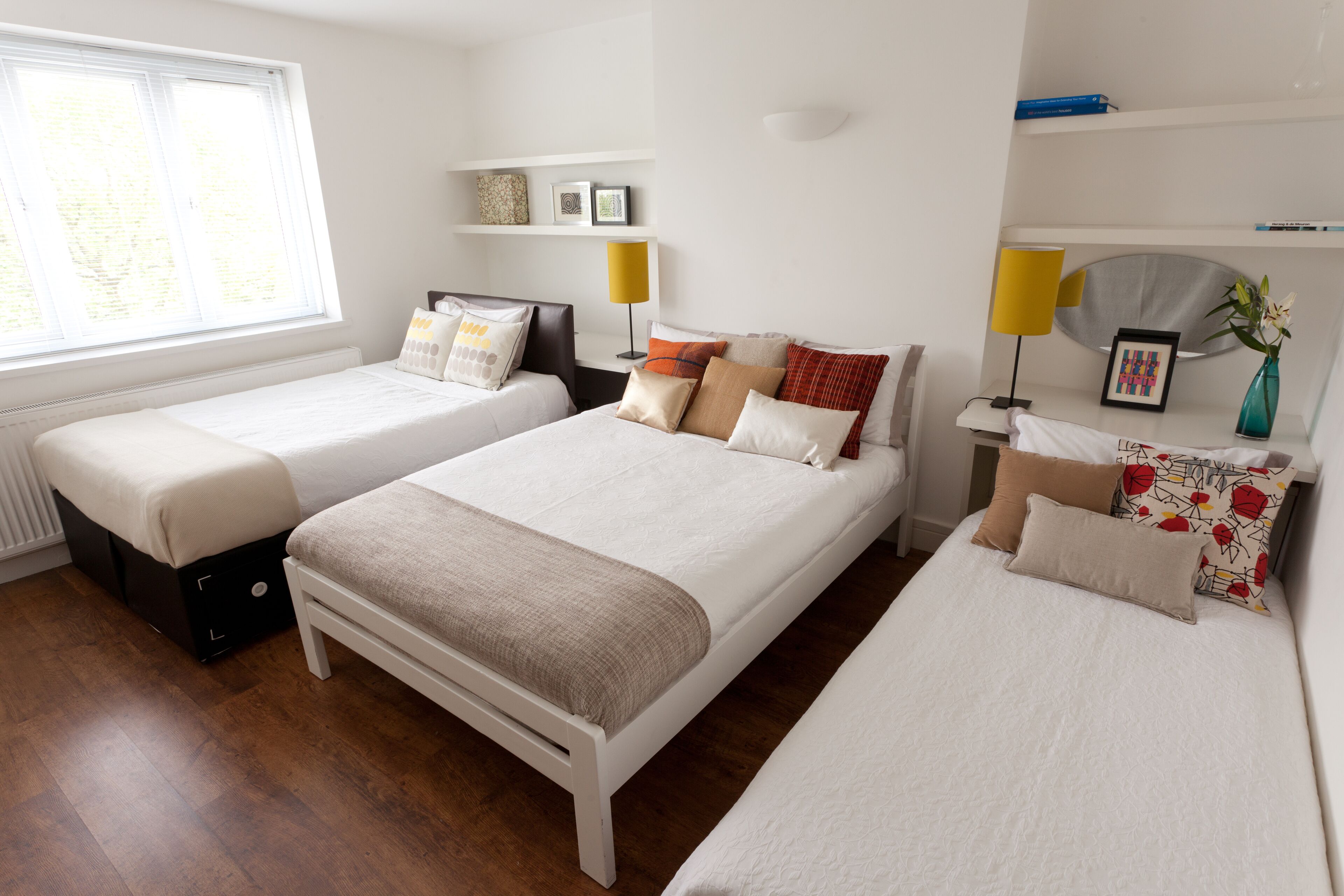 Une chambre d’hôtel avec deux lits simples, une table de chevet avec une lampe, une petite étagère avec des objets décoratifs et une fenêtre avec des stores.