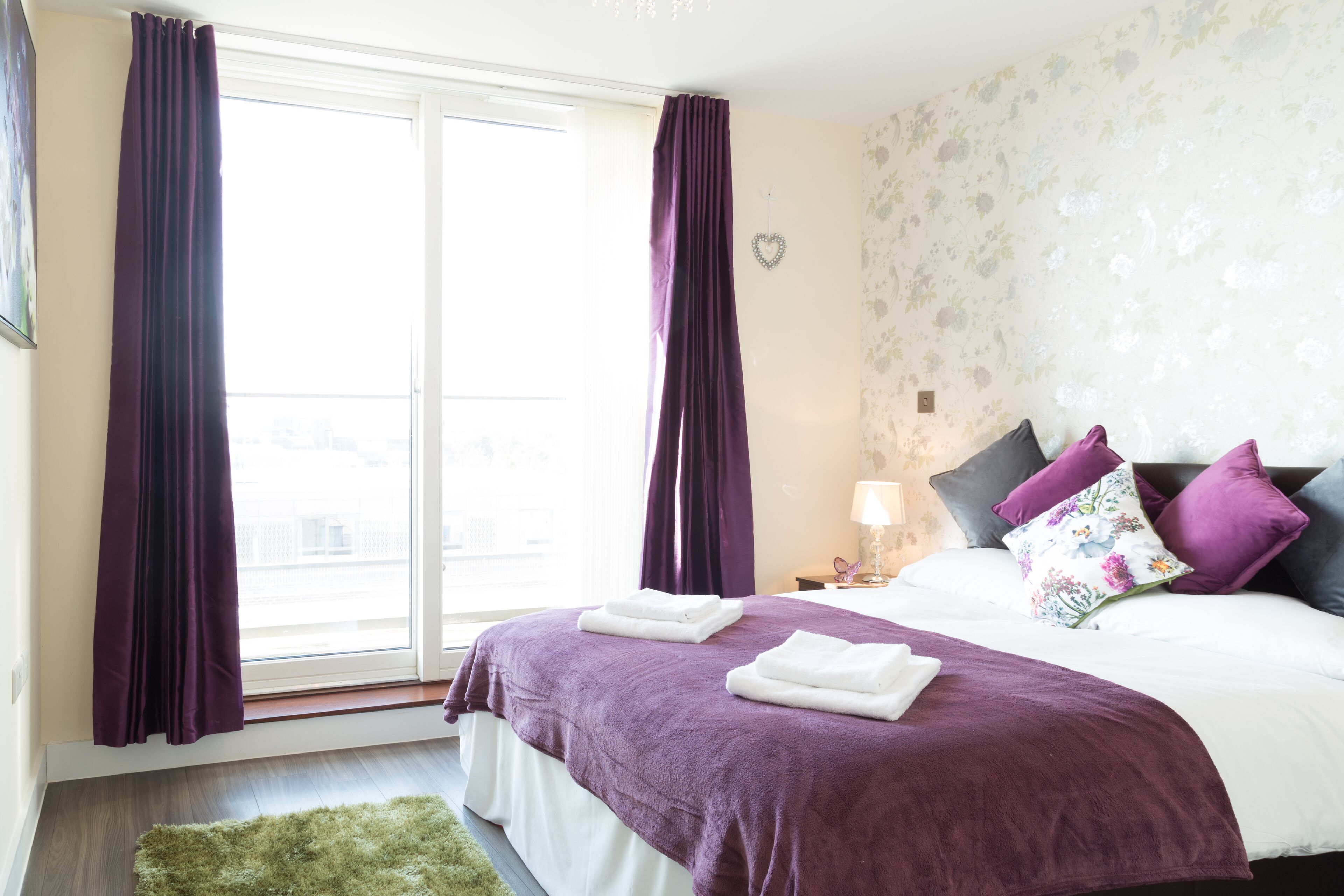 A hotel room with a large window, a bed with purple bedding, and a green rug.