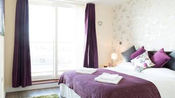 A hotel room with a large window, a bed with purple bedding, and a green rug.