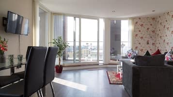 A modern living room with a large sliding glass door leading to a balcony, a flat-screen TV mounted on the wall, and a grey sofa with floral cushions.
