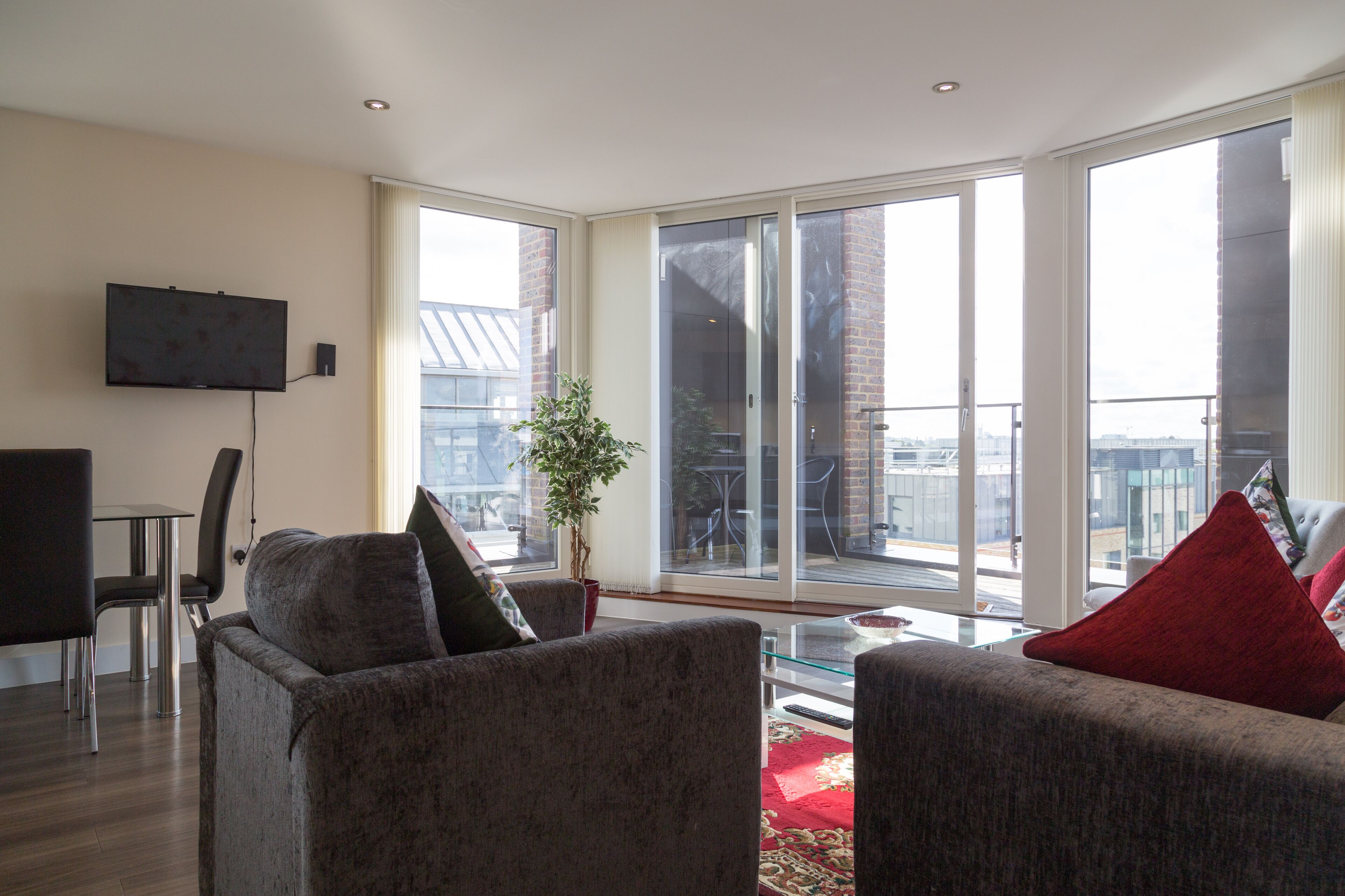 A modern living room with a flat-screen TV mounted on the wall, a dining area with a table and chairs, and large windows offering a view of the city.