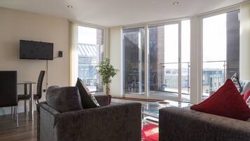 A modern living room with a flat-screen TV mounted on the wall, a dining area with a table and chairs, and large windows offering a view of the city.