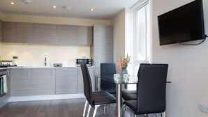 A modern kitchen with grey cabinets, a glass dining table, black chairs, and a flat-screen TV mounted on the wall. - Exquisite Modern Station Penthouse with Balcony, Parking & Sleeps 6 (Cambridge)