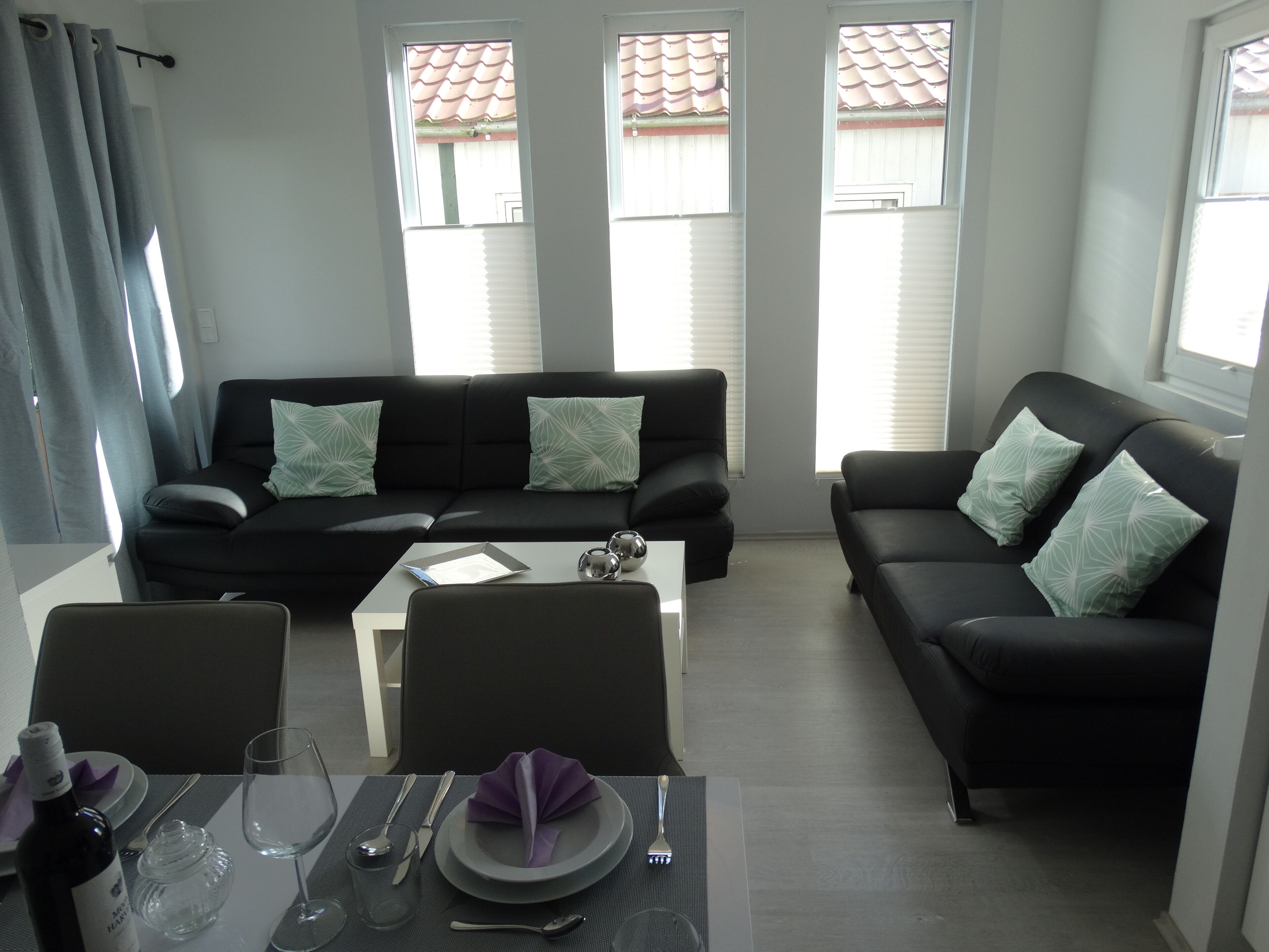 Un salon moderne avec un canapé noir, deux fauteuils assortis, une table basse et un coin repas avec une table dressée pour deux personnes.