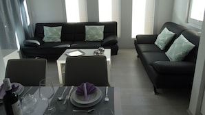 Een moderne woonkamer met een zwarte bank, twee bijpassende fauteuils, een salontafel en een eethoek met een tafel gedekt voor twee personen.