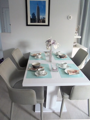 Un coin repas avec une table dressée pour deux personnes, agrémenté d’un vase de fleurs, d’une vue de la ville encadrée et d’une chaise au design moderne.