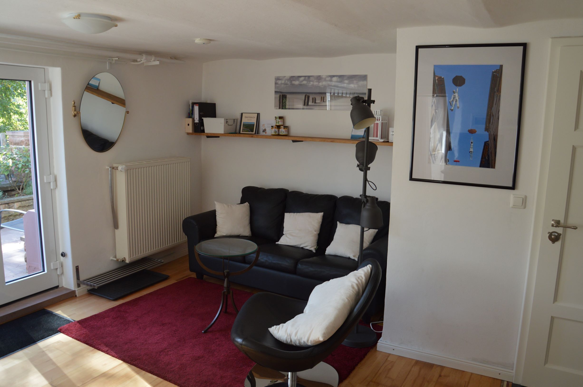 A living room with a black leather sofa, a round mirror, a framed picture, and a red rug.