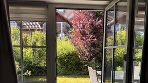 Huone, jossa on liukuovet, jotka johtavat ulkoterasille, jossa on pöytä ja tuoleja. Ovella on etumatto, jossa lukee ”KOTI”, ja huoneesta on näkymä puutarhaan ja naapuritaloihin.