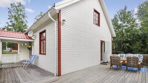 Uma casa branca com telhado vermelho, um deck com mesa e cadeiras, e uma cadeira dobrável azul.