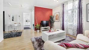 Uma sala de estar moderna com um sofá branco, uma mesa de centro e uma TV. Há uma parede em vermelho, uma planta e uma janela com cortinas.
