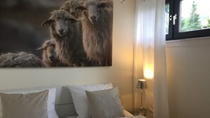 A bedroom with a bed, pillows, a nightstand with a lamp, a wall-mounted painting of sheep, and a window with a view of trees. - Romantic apartment with fantastic views, WiFi free (Winterberg)