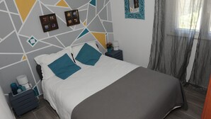 Ein ordentlich bezogenes Bett mit einer weiß-grauen Bettdecke, zwei türkisfarbenen Kissen und einer dunkelgrauen Tagesdecke. Ein kleiner Nachttisch mit Lampe, einer Vase mit Blumen und einem gerahmten Foto. An der Wand ist ein geometrisches Muster mit türkisfarbene Akzenten zu sehen.
