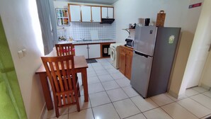 A compact kitchen with a dining table and chairs, a washing machine, a refrigerator, and tiled flooring. - Manava Tahara'a home stay (Mahina)