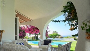 Un área de estar al aire libre techada con mobiliario de mimbre, una piscina y vistas al mar.