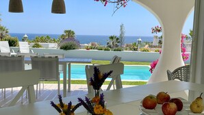 Una mesa con una bandeja de frutas y arreglos florales, con vista a una piscina y el océano.