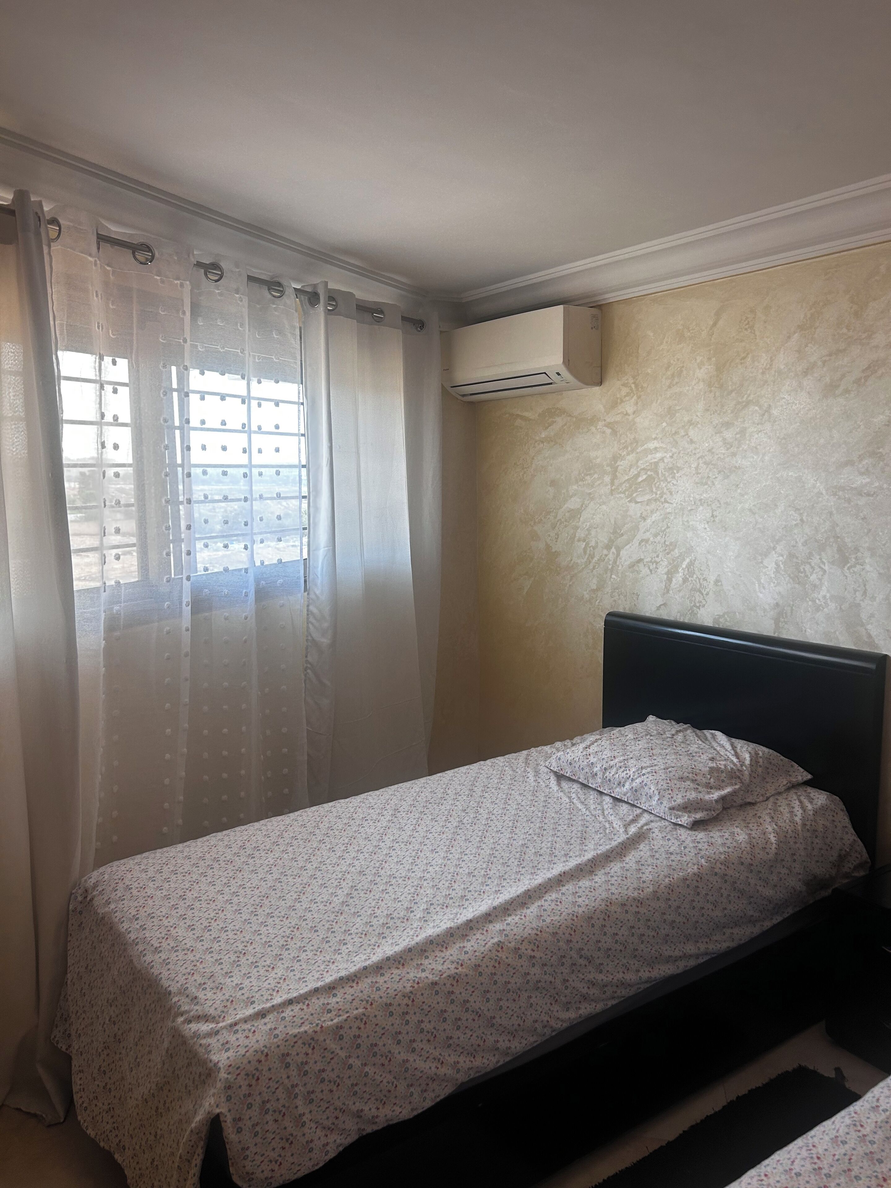 Une petite chambre avec un lit simple, un lit sans fioritures, une fenêtre avec des rideaux légers et un appareil de climatisation fixé au mur.