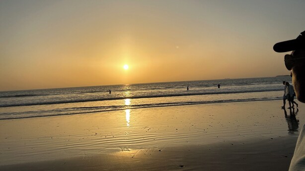 Une scène de plage avec le soleil se couchant sur l’océan, des personnes se promenant le long du rivage et un ciel dégagé.