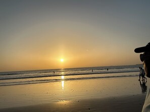 Une scène de plage avec le soleil se couchant sur l’océan, des personnes se promenant le long du rivage et un ciel dégagé.