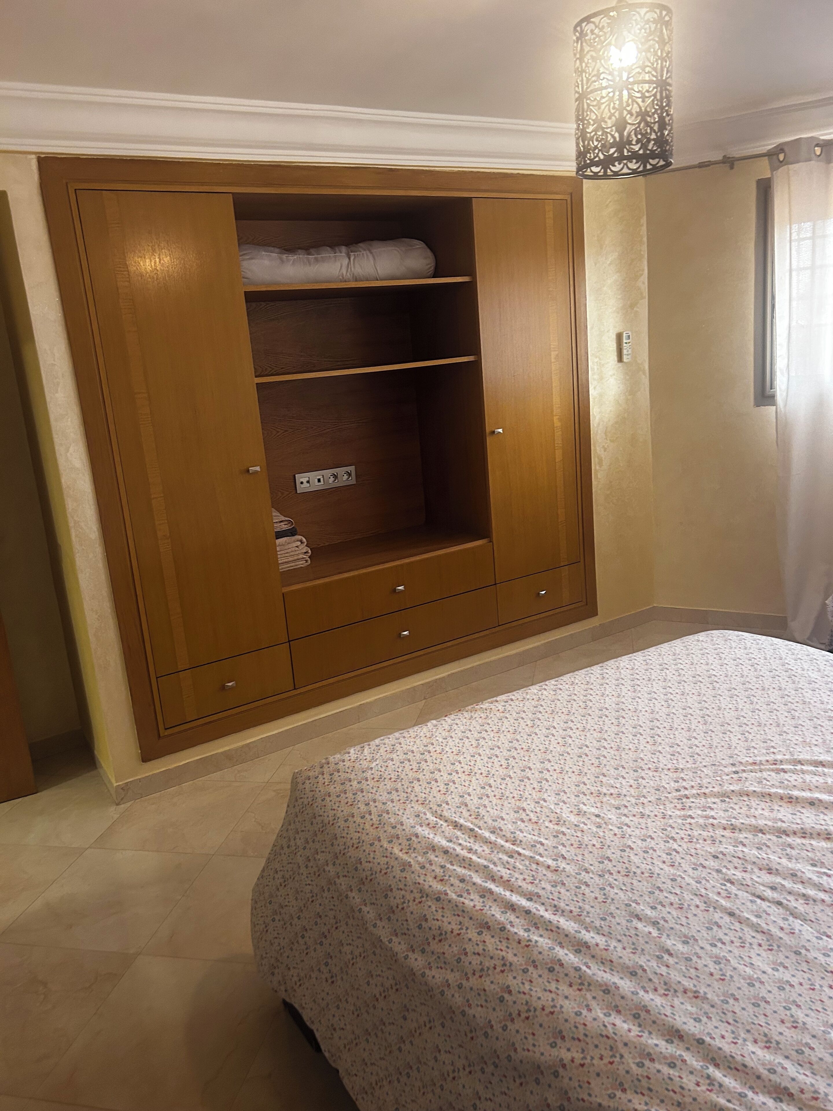 A bedroom with a large wooden wardrobe, a bed with a floral bedspread, and a hanging light fixture.