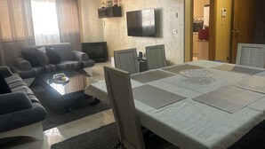 Un salon avec un canapé, une table basse et un téléviseur. On aperçoit une table à manger avec des chaises, ainsi qu’une cuisine à l’arrière-plan.