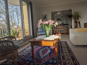 Un salon doté d’une grande fenêtre, d’une table basse en bois sur laquelle est posé un vase de fleurs, d’un tapis à motifs, d’un fauteuil en osier et d’un canapé blanc.