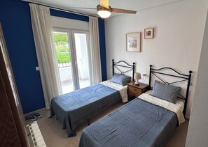 Un dormitorio con dos camas, un ventilador de techo, una mesita de noche con lámpara y una ventana con cortinas.